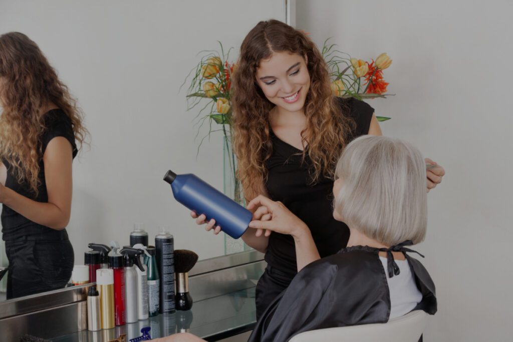 Una giovane parrucchiera sorridente con i capelli ricci tiene un flacone blu di prodotto per capelli mentre consiglia una donna anziana dai capelli argentati seduta su una poltrona in un salone.