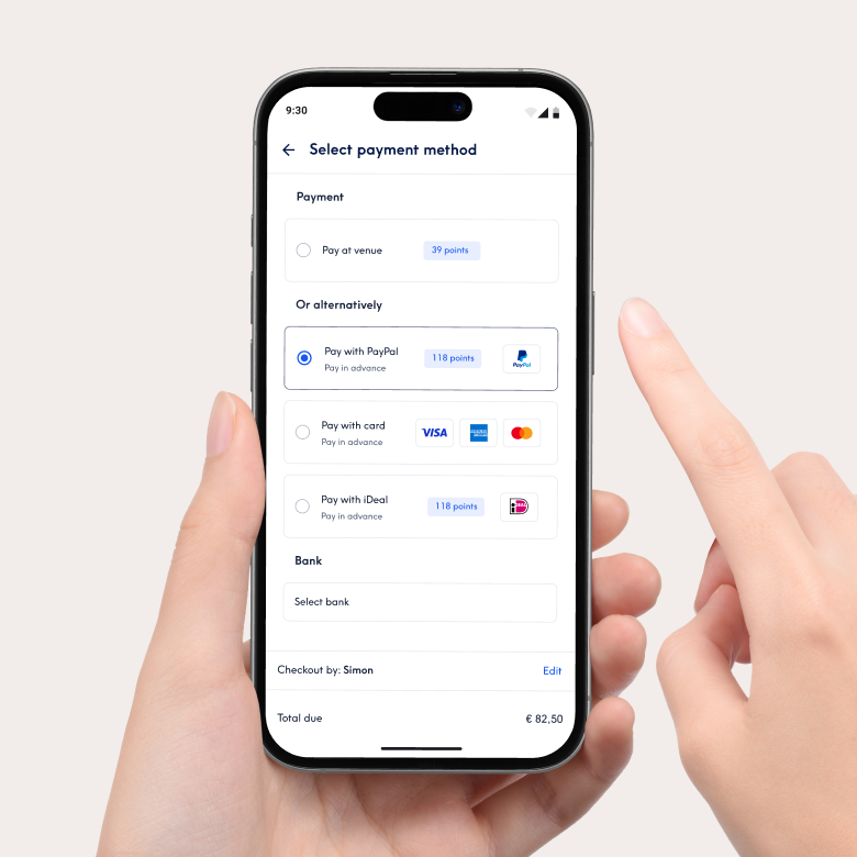 Primo piano di uno smartphone che mostra l'interfaccia di pagamento dell'app Treatwell per la selezione del metodo di pagamento. Tra le opzioni ci sono il pagamento in salone o il pagamento anticipato con PayPal, carta di credito o iDeal. Una mano tiene il telefono e un dita ne indica lo schermo.