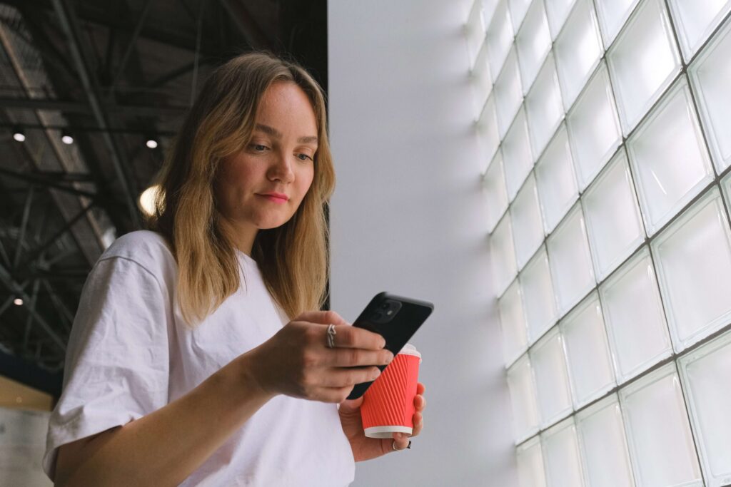 Inquadratura dal basso di una giovane donna con i capelli biondi e una t-shirt bianca, che guarda il suo smartphone nero mentre tiene in mano una tazza di caffè da asporto rossa. Si trova accanto a una grande parete di vetromattone.