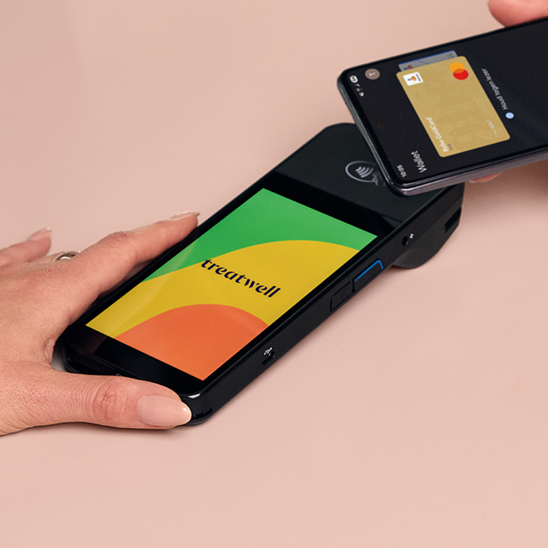 Una mano tiene un terminale di pagamento Treatwell nero mentre un cliente avvicina lo smartphone con wallet digitale per pagare in modalità contactless.